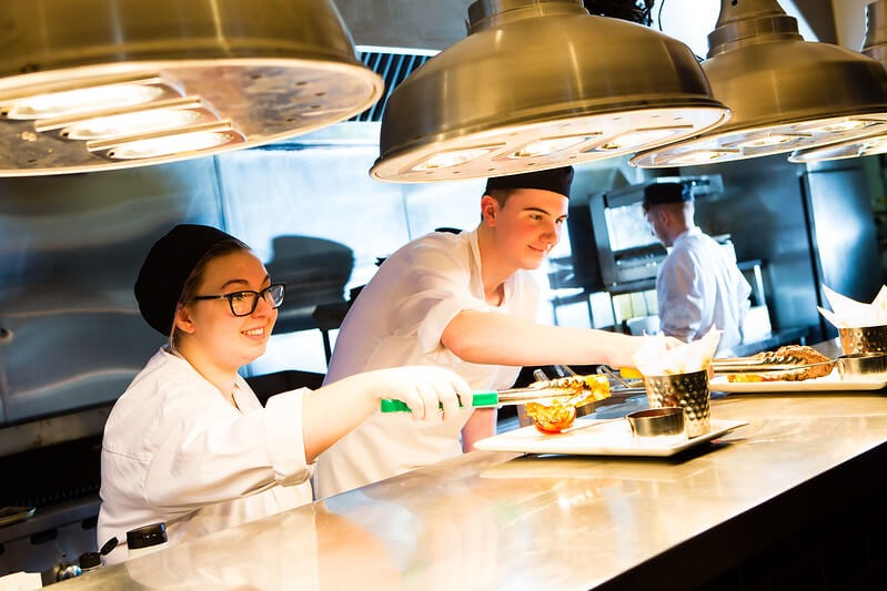 girl and boy wearing chef whites plating up food in a commercial kitchen